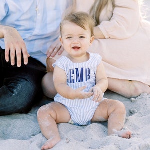 May include: A baby wearing a blue and white striped onesie with the letters "EMB" embroidered on the front. The baby is sitting in the sand on a beach.