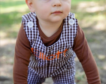 Puede incluir: Un niño pequeño con un mono de pana marrón y blanco con el nombre "Elliot" bordado en naranja. El niño está de pie sobre una superficie de madera y mirando hacia arriba.