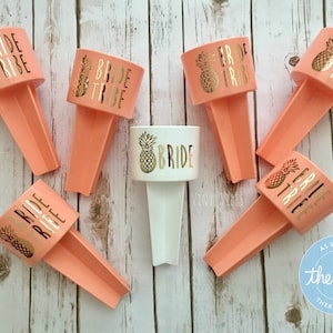 May include: Six peach-colored plastic drink holders with a pineapple design and the text "Bride Tribe" in gold lettering. One white plastic drink holder with a pineapple design and the text "Bride" in gold lettering.