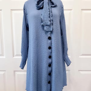 May include: A light blue textured long-sleeve dress with a ruffled collar and bow. The dress features a row of black buttons down the front and a slightly flared skirt. The fabric has a subtle, crinkled appearance.