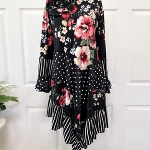 May include: A black long-sleeve dress with a floral print featuring large pink flowers and white polka dots. The dress has a ruffled hem and sleeve cuffs, with a striped pattern on the lower portion. A large fabric flower adorns the shoulder.