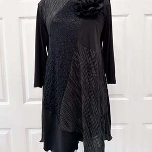 May include: Black tunic top with a large black flower embellishment on the neckline. The top is made of different fabrics, including a textured fabric, a smooth fabric, and a sheer fabric. The top has a long sleeve and a loose fit.