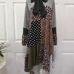 May include: A black and white striped, polka dot, floral, and leopard print dress with a black bow tie. The dress has a high neck and long sleeves. The hemline is asymmetrical and falls to the knee.