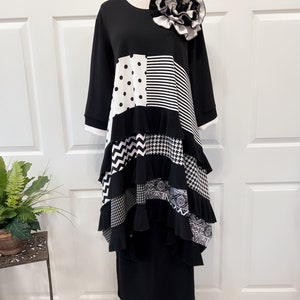 May include: Black and white patchwork dress with a ruffled hem and a large black and white flower accent. The dress has a long sleeve and a round neckline.