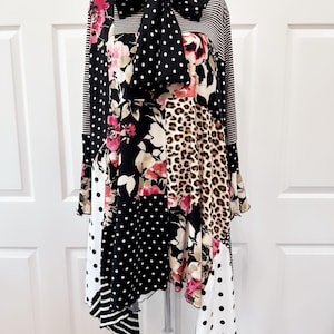 May include: A patchwork dress with a variety of patterns, including polka dots, floral prints, and leopard print. The dress features long sleeves, a bow at the neckline, and a mix of black, white, and pink colors. The dress is displayed on a mannequin.