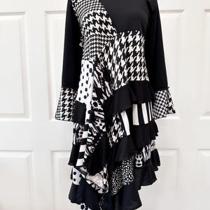 Könnte beinhalten: Schwarz-weißes Patchworkkleid mit einem Rüschen-Saum. Das Kleid hat lange Ärmel und einen hohen Kragen. Der Stoff ist eine Mischung aus verschiedenen Mustern, darunter Hahnentritt, Punkte und Streifen.