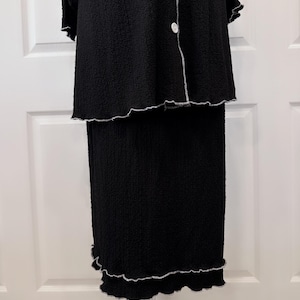 May include: A black two-piece outfit with a long-sleeved top and a skirt. The top has a button-up front and a ruffled hem. The skirt has a ruffled hem and is slightly longer than the top.