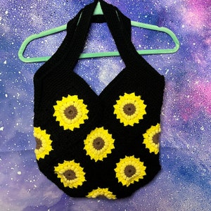Puede incluir: Una bolsa de mano de crochet negra con un diseño de correa halter. La bolsa está decorada con girasoles de crochet amarillos y marrones.
