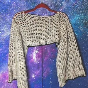 May include: A beige, open-weave crochet top with long, flared sleeves. The cropped sweater has a black trim along the bottom edge. The garment is hanging on a red hanger against a blue and purple galaxy-themed background.