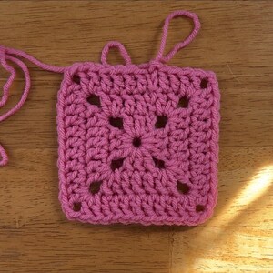 May include: A pink crochet square with a central flower design. The square has a textured, crocheted surface and is made with a single color of yarn.