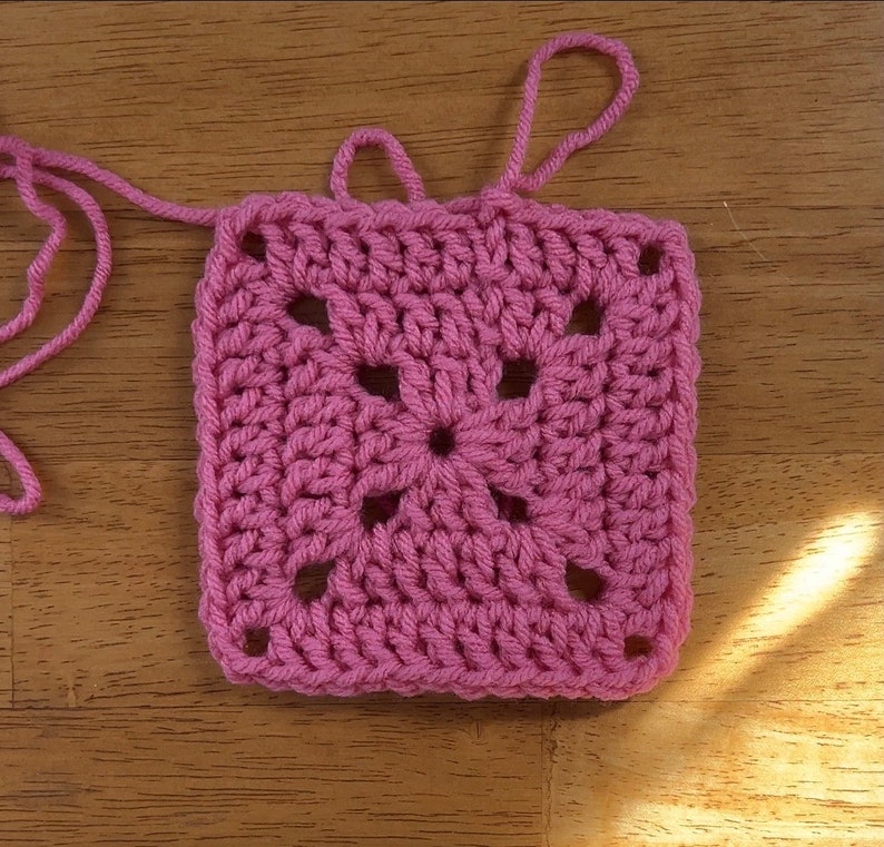 May include: A pink crochet square with a central flower design. The square has a textured, crocheted surface and is made with a single color of yarn.