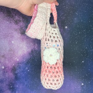 Puede incluir: Un porta botellas de crochet rosa y blanco con una flor de crochet blanca con un centro brillante.