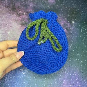 May include: A handmade, blue crochet drawstring pouch with a green drawstring. The pouch is held in a hand against a galaxy background. The drawstring is secured with two small silver beads. The pouch is approximately 5 inches wide.