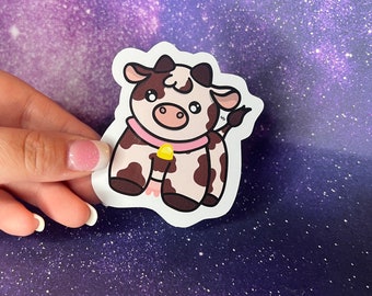 May include: A die-cut sticker featuring a cartoon cow with brown and white spots. The cow has a pink collar and a yellow object. The sticker is held against a purple and blue starry background.
