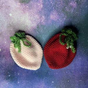 Puede incluir: Dos bolsitas de punto de ganchillo con forma de fresa, una rosa y otra roja, con hojas de ganchillo verdes en la parte superior.