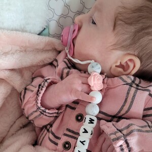 Peut inclure: Une attache-sucette compos&eacute;e d'une cha&icirc;ne de perles rondes en silicone blanches, grises &agrave; motif marbr&eacute; et rose p&acirc;le, avec une perle en forme de rose et des perles carr&eacute;es blanches formant le pr&eacute;nom MAYA, fix&eacute;e &agrave; une pince m&eacute;tallique et une sucette rose.