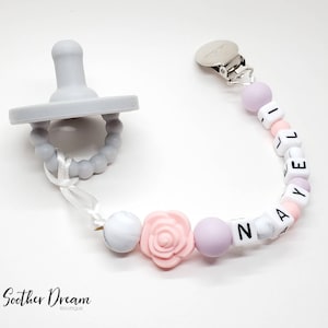 Peut inclure: Une sucette en silicone gris clair attach&eacute;e &agrave; une attache-sucette perl&eacute;e avec des perles en silicone blanches, rose clair et lavande. Les perles forment le nom NAYELI et incluent une perle en forme de rose rose clair, avec un clip en m&eacute;tal argent&eacute;.