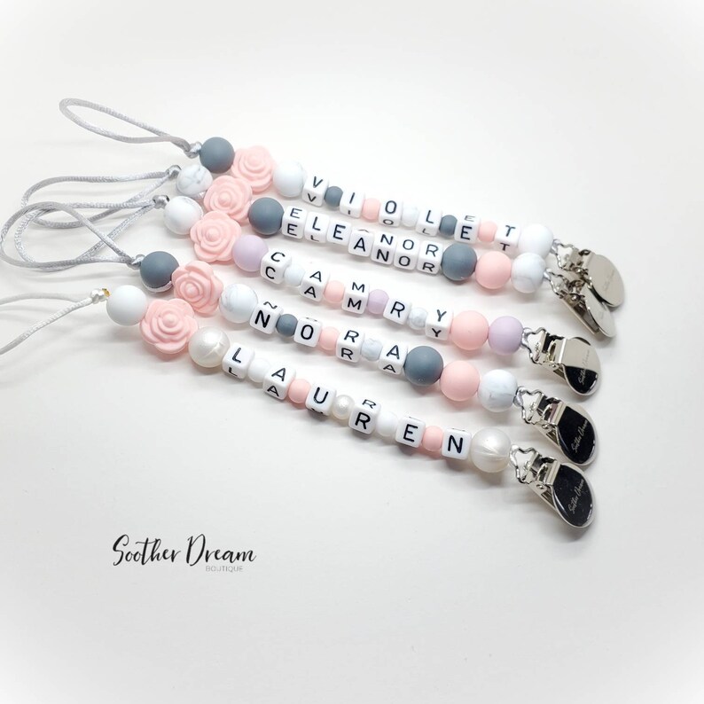 Peut inclure: Quatre attaches-sucettes personnalis&eacute;es avec des perles en silicone blanches, grises et rose clair, incluant des perles en forme de rose et des cubes avec les lettres formant les pr&eacute;noms Violet, Eleanor, Camry, Nora et Lauren. Clip en m&eacute;tal argent&eacute;.