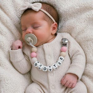 May include: A beige dummy clip featuring round silicone beads in shades of pink, beige, and white, with white letter beads spelling the name SCOTTIE. The clip has a metal fastener and is attached to a beige dummy and a matching headband.