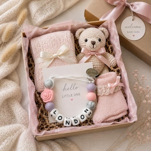 Peut inclure: Un coffret cadeau contenant une couverture tricot&eacute;e rose p&acirc;le avec un ruban, un ours en crochet avec un n&oelig;ud rose, une paire de chaussettes roses avec dentelle et une attache-sucette avec des perles &eacute;pelant LONDON et une carte hello LITTLE ONE.