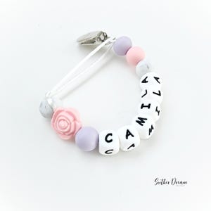 Peut inclure: Une attache-sucette avec un clip en m&eacute;tal argent&eacute;, un cordon blanc et une s&eacute;rie de perles. Les perles incluent des sph&egrave;res lavande, rose et blanc marbr&eacute;, une perle en forme de rose rose et des cubes blancs &eacute;pelant le nom CAMILA.