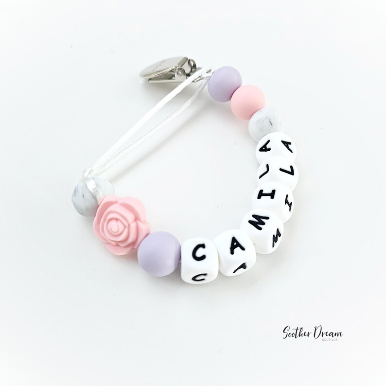 Peut inclure: Une attache-sucette avec un clip en m&eacute;tal argent&eacute;, un cordon blanc et une s&eacute;rie de perles. Les perles incluent des sph&egrave;res lavande, rose et blanc marbr&eacute;, une perle en forme de rose rose et des cubes blancs &eacute;pelant le nom CAMILA.
