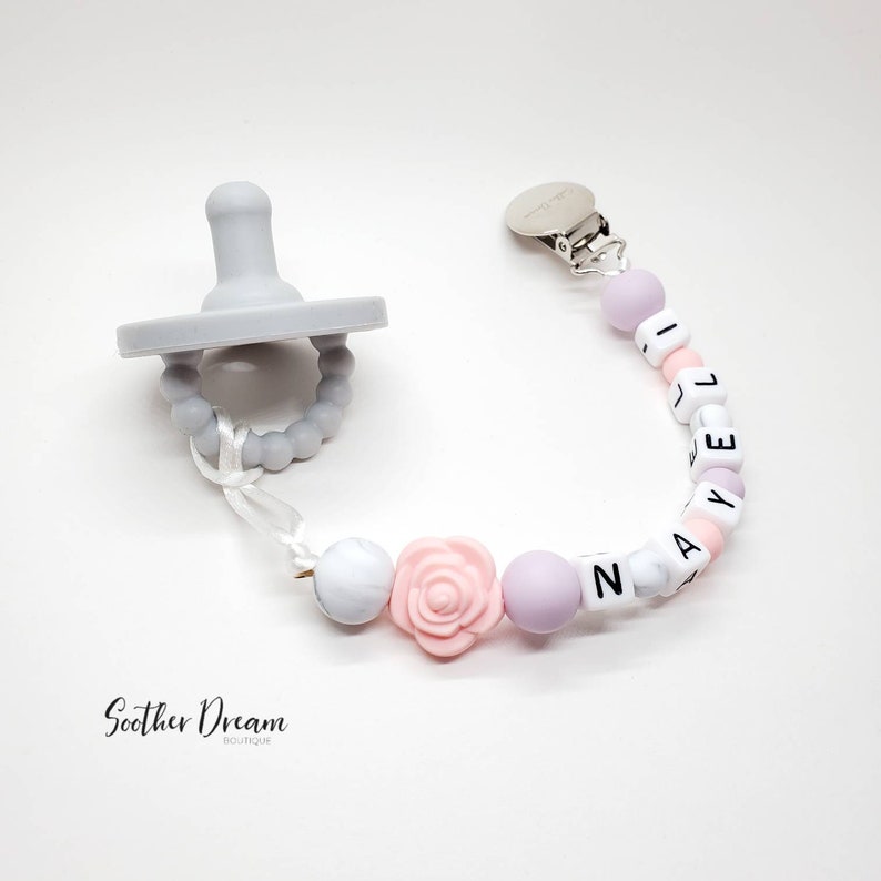 Peut inclure: Une sucette en silicone gris clair attach&eacute;e &agrave; une attache-sucette perl&eacute;e avec des perles en silicone blanches, rose clair et lavande. Les perles forment le nom NAYELI et incluent une perle en forme de rose rose clair, avec un clip en m&eacute;tal argent&eacute;.