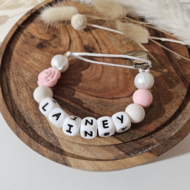Peut inclure: Une attache-sucette compos&eacute;e de perles blanches et roses, incluant une perle en forme de rose et des perles cubiques blanches avec le pr&eacute;nom LAINEY imprim&eacute; en noir. Les perles sont enfil&eacute;es sur un cordon blanc fix&eacute; &agrave; une pince m&eacute;tallique argent&eacute;e.