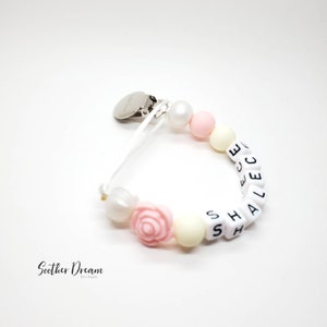 Peut inclure: Une attache-sucette avec un clip en m&eacute;tal argent&eacute; fix&eacute; &agrave; un ruban blanc. Le cordon comprend des perles rondes blanches et jaune p&acirc;le, une perle en forme de rose rose et des perles cubiques blanches avec des lettres noires formant le nom SHALECE.