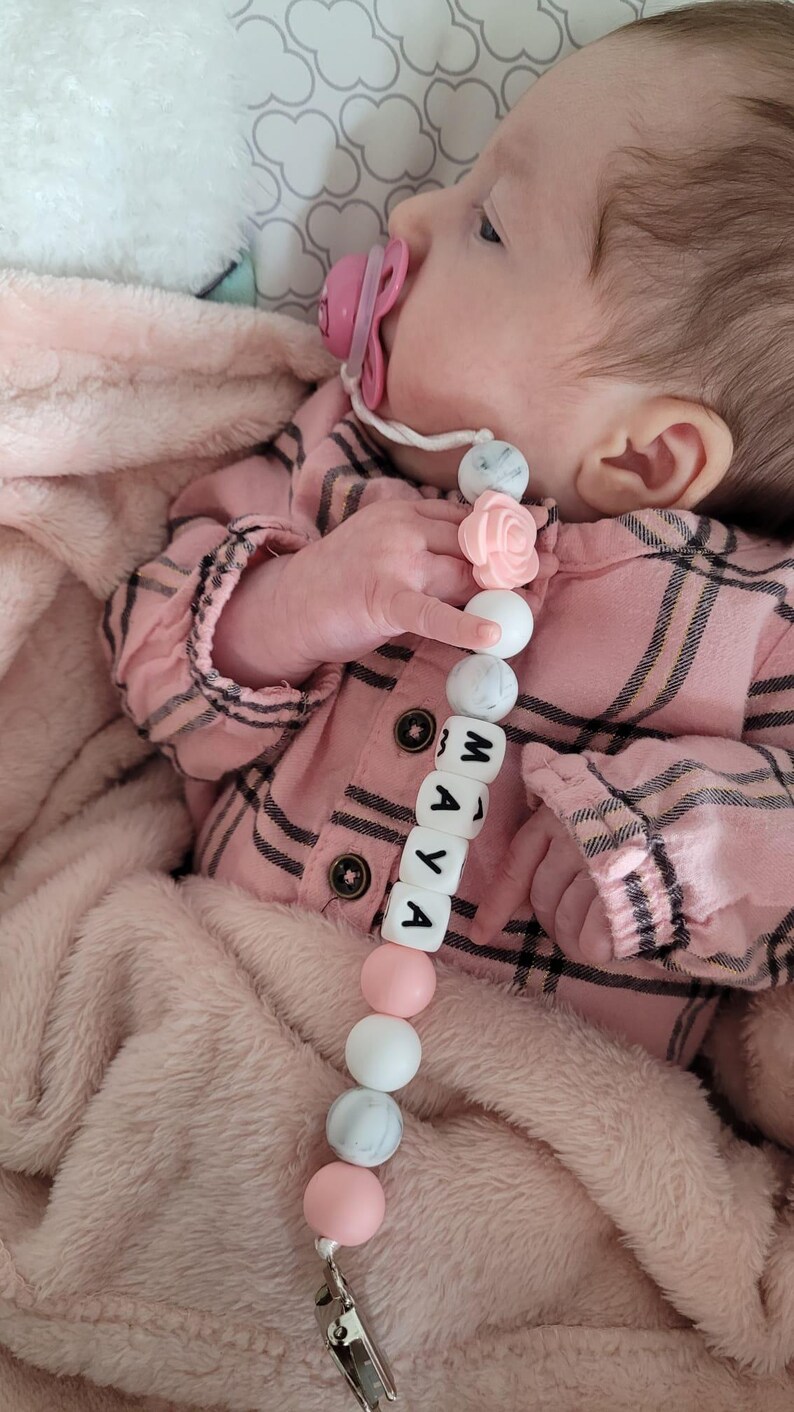 Peut inclure: Une attache-sucette compos&eacute;e d'une cha&icirc;ne de perles rondes en silicone blanches, grises &agrave; motif marbr&eacute; et rose p&acirc;le, avec une perle en forme de rose et des perles carr&eacute;es blanches formant le pr&eacute;nom MAYA, fix&eacute;e &agrave; une pince m&eacute;tallique et une sucette rose.