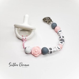 Peut inclure: Une sucette blanche attach&eacute;e &agrave; une attache-sucette perl&eacute;e. L'attache comporte des perles en silicone rondes grises et roses, des perles lettres blanches formant le nom BLAKE et une perle en forme de rose rose. Un clip m&eacute;tallique est fix&eacute;.