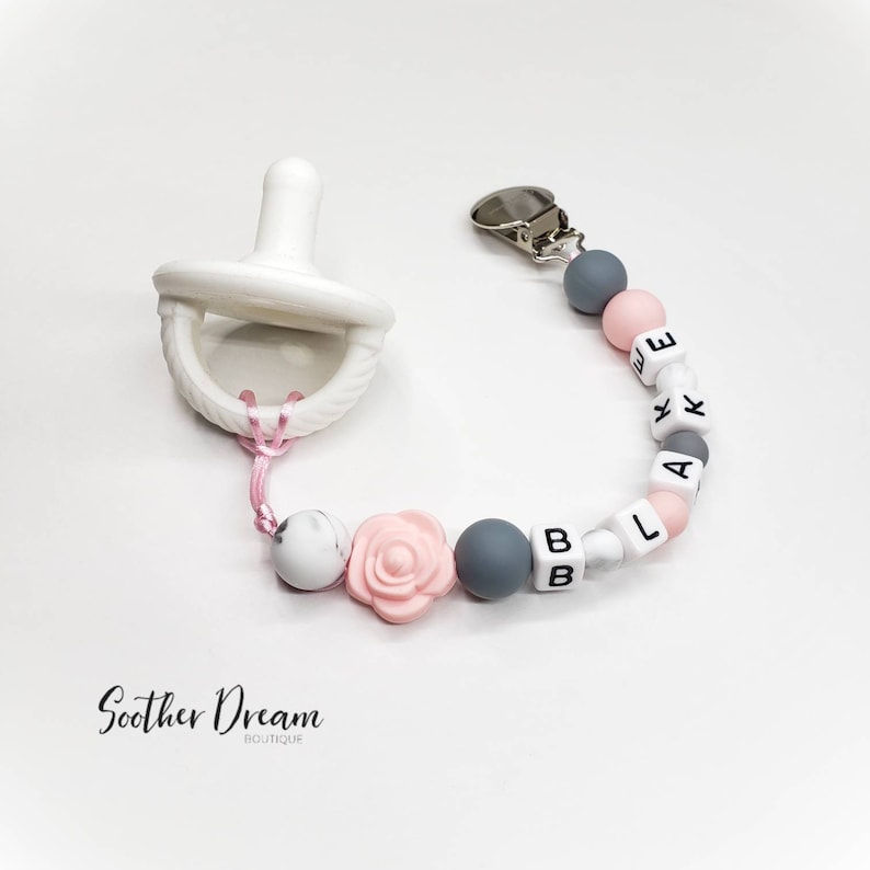 Peut inclure: Une sucette blanche attach&eacute;e &agrave; une attache-sucette perl&eacute;e. L'attache comporte des perles en silicone rondes grises et roses, des perles lettres blanches formant le nom BLAKE et une perle en forme de rose rose. Un clip m&eacute;tallique est fix&eacute;.