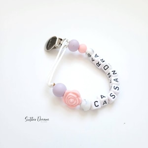 Peut inclure: Une attache-sucette avec un clip en m&eacute;tal argent&eacute;, un cordon blanc et une s&eacute;rie de perles comprenant des sph&egrave;res lavande, roses et blanches &agrave; motif marbr&eacute;, une perle en forme de rose rose et des perles carr&eacute;es blanches &eacute;pelant CASSANDRA.