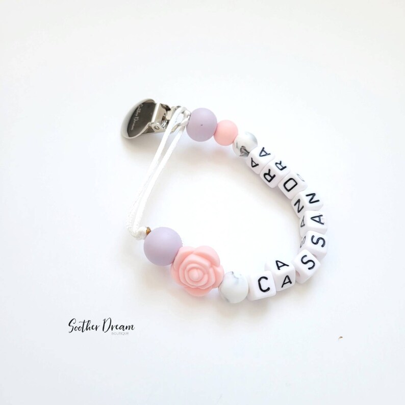 Peut inclure: Une attache-sucette avec un clip en m&eacute;tal argent&eacute;, un cordon blanc et une s&eacute;rie de perles comprenant des sph&egrave;res lavande, roses et blanches &agrave; motif marbr&eacute;, une perle en forme de rose rose et des perles carr&eacute;es blanches &eacute;pelant CASSANDRA.