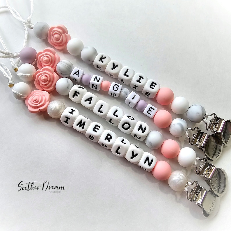 Peut inclure: Quatre attaches-sucettes compos&eacute;es de perles en silicone blanches, roses et &agrave; motif marbr&eacute;, incluant une perle en forme de rose et des cubes blancs portant les noms KYLIE, ANGIE, FALLON et IMERLYN, avec des clips m&eacute;talliques argent&eacute;s et cordons.