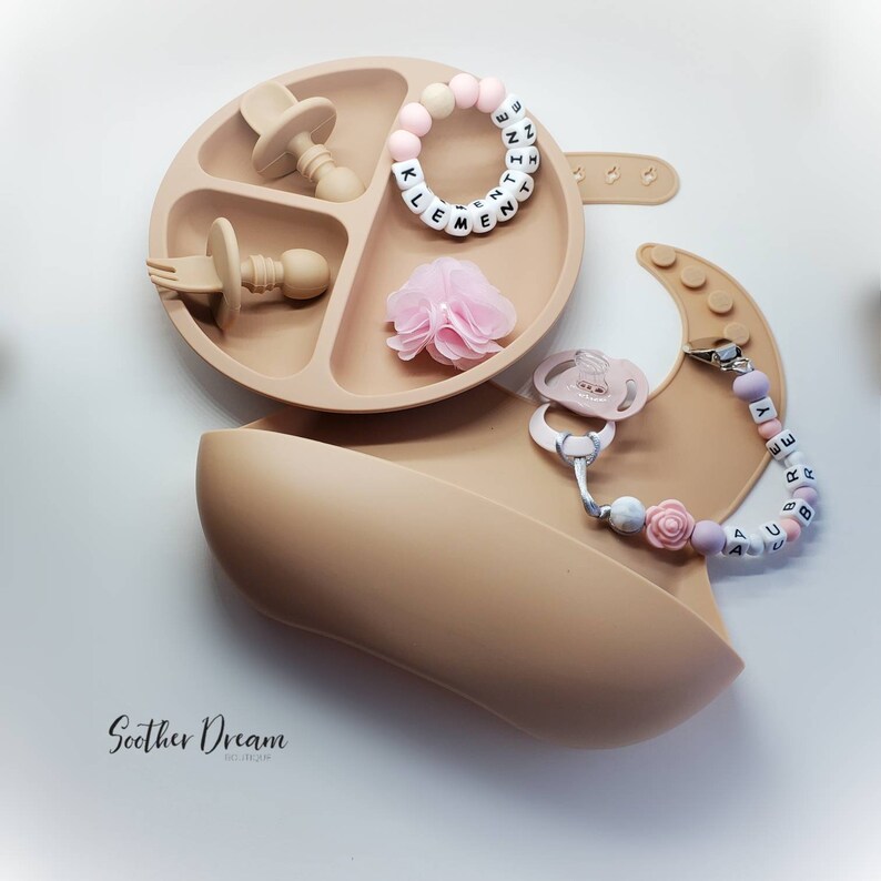 Peut inclure: Une assiette &agrave; ventouse en silicone beige &agrave; trois compartiments contenant une cuill&egrave;re et une fourchette assorties, un anneau de perles personnalis&eacute; au nom de KLEMENTINE, une fleur en tissu rose, un bavoir en silicone beige et une sucette.