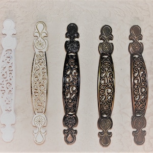 May include: Five ornate metal drawer pulls in different finishes: white, gold, black, bronze, and brass. Each pull features a decorative, intricate design with a floral motif.