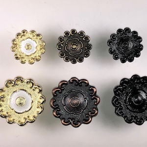 May include: Six decorative cabinet knobs with a floral design. The knobs are in three different colours: gold, black, and bronze. The knobs have a raised centre with a smaller circle in the centre.