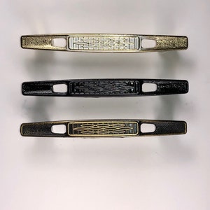 May include: Three cabinet door pulls in different finishes. The top pull is gold with a white design, the middle pull is black, and the bottom pull is black with a gold design.
