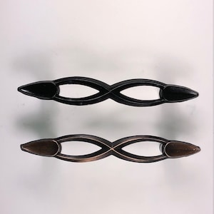 Könnte beinhalten: Zwei längliche Schrankgriffe mit einem Design aus zwei ineinander verschlungenen Schlaufen. Ein Griff hat eine einfarbige, mattschwarze Oberfläche, während der andere eine dunkelbronzene Oberfläche mit kupferfarbenen Akzenten an den Kanten aufweist.