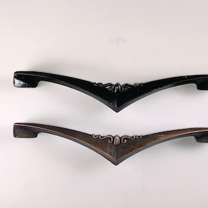May include: Two cabinet pulls, one black and one copper, with a decorative design in the center. The pulls are shaped like a curved arch with a pointed end.