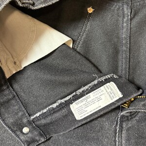 May include: Close-up of a black denim Wrangler jeans label with the text "Wrangler 12MWZMS 11/12X34" and care instructions. The label is attached to the inside of the jeans.