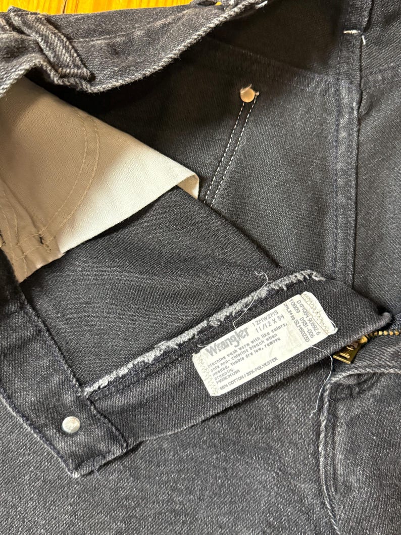 May include: Close-up of a black denim Wrangler jeans label with the text "Wrangler 12MWZMS 11/12X34" and care instructions. The label is attached to the inside of the jeans.