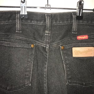 May include: Black denim jeans with a brown leather Wrangler patch on the back pocket. The jeans have a classic five-pocket style and a straight leg.