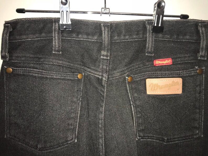 May include: Black denim jeans with a brown leather Wrangler patch on the back pocket. The jeans have a classic five-pocket style and a straight leg.