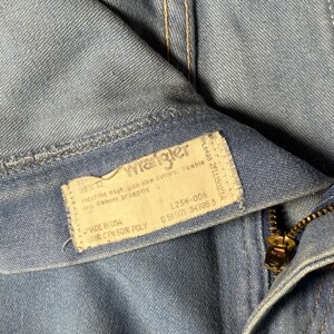May include: Close-up of a blue denim Wrangler jeans label with the text "Wrangler" and care instructions. The label also includes the size, style number, and fabric content. The jeans have a zipper fly and a metal zipper pull.