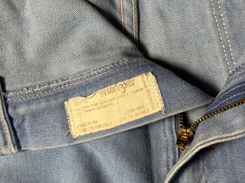 May include: Close-up of a blue denim Wrangler jeans label with the text "Wrangler" and care instructions. The label also includes the size, style number, and fabric content. The jeans have a zipper fly and a metal zipper pull.