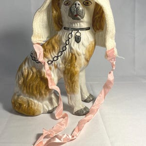 May include: A porcelain figurine of a dog wearing a white knitted hat with a pink ribbon tied around its neck. The dog is sitting with its head tilted slightly to the side.
