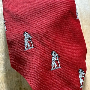 Puede incluir: Una corbata roja con un patrón repetido de pequeñas figuras blancas y grises. La corbata tiene una superficie texturizada y está en ángulo, mostrando el tejido de la tela. Las figuras parecen ser ilustraciones de una persona sosteniendo un bastón.