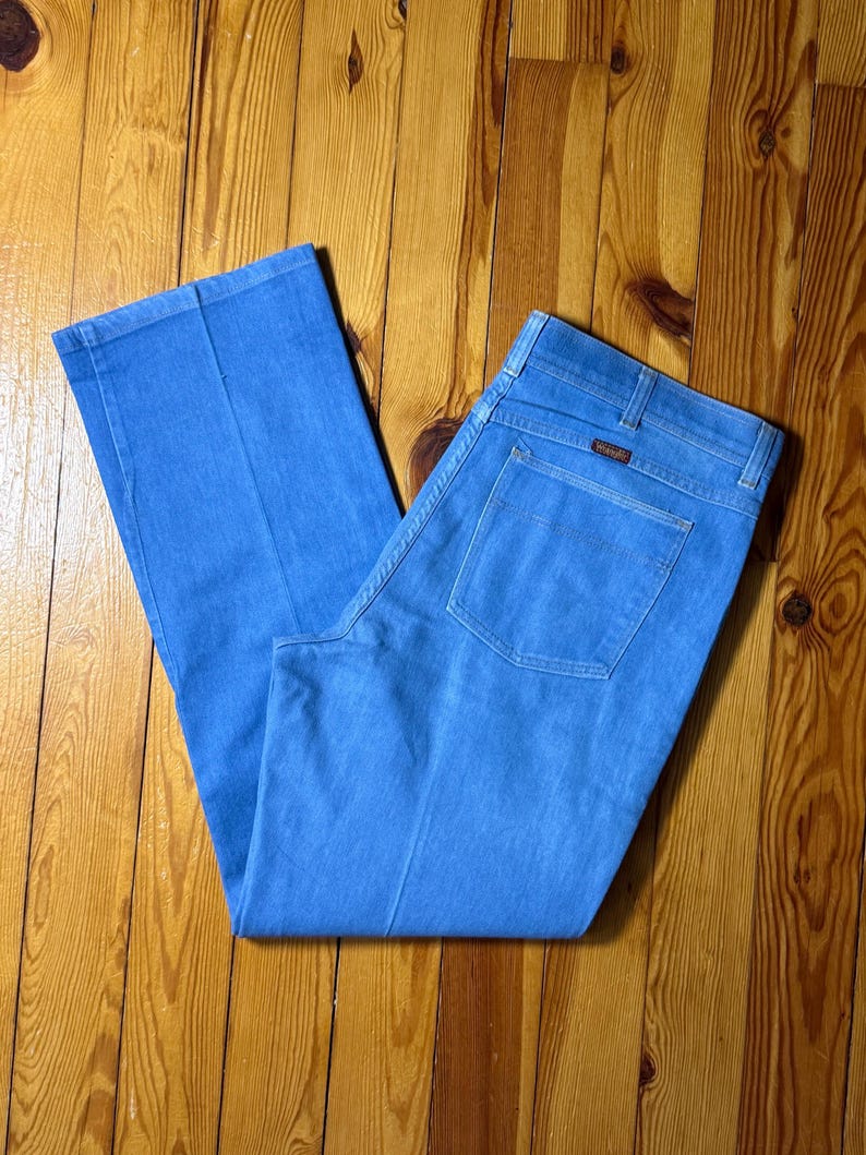 May include: A pair of light blue denim jeans with a straight leg and a classic five-pocket design. The jeans have a brown leather patch with the Wrangler logo on the back.